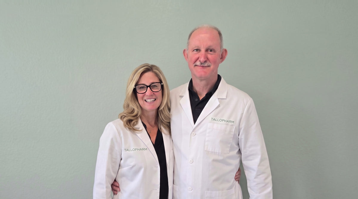 A man and a woman wearing white lab coats with 'TALLOPHARMA' embroidered on the chest, standing against a light sage green wall.