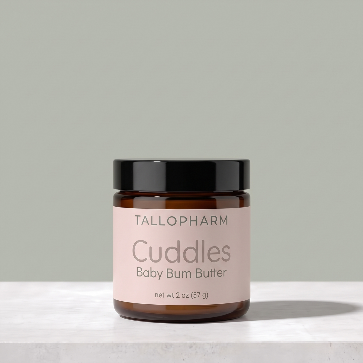 A jar of TALLOPHARM Cuddles Baby Bum Butter, featuring a black lid and a pale pink label, set on a light gray marble surface against a solid sage green background.