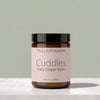 A jar of Tallopharm Cuddles Daily Diaper Balm, featuring an amber glass jar, a black lid, and a pale pink label, set on a light marble surface against a muted sage green background.