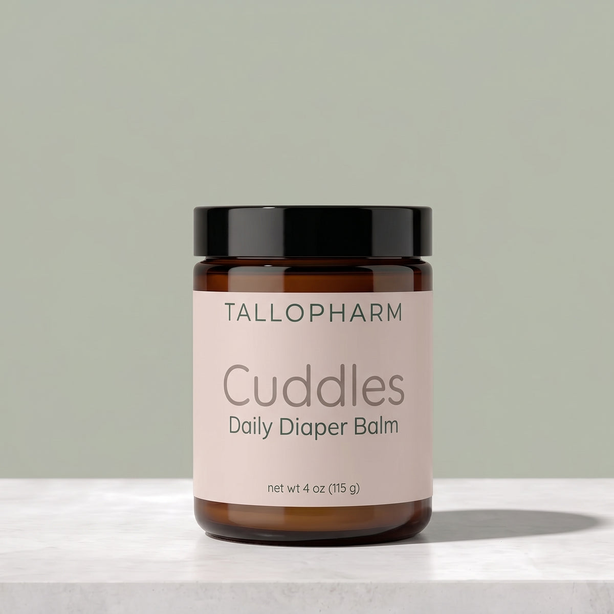 A jar of Tallopharm Cuddles Daily Diaper Balm, featuring an amber glass jar, a black lid, and a pale pink label, set on a light marble surface against a muted sage green background.