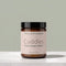 A jar of Tallopharm Cuddles Daily Diaper Balm, featuring an amber glass jar, a black lid, and a pale pink label, set on a light marble surface against a muted sage green background.