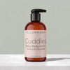A bottle of Tallopharm Cuddles Baby Body Lotion with a black pump and a pale pink label, set on a light marble surface against a soft gray-green background.