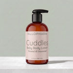 A bottle of Tallopharm Cuddles Baby Body Lotion with a black pump and a pale pink label, set on a light marble surface against a soft gray-green background.