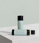 Two lip balm tubes from TalloPharm, one standing upright and one lying horizontally, both in a pale mint green color with black caps, set on a white marble surface against a matching pale green background. Berry flavour.