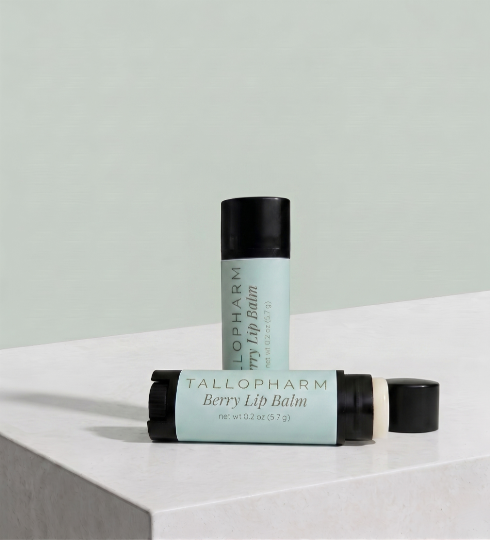 Two lip balm tubes from TalloPharm, one standing upright and one lying horizontally, both in a pale mint green color with black caps, set on a white marble surface against a matching pale green background. Berry flavour.