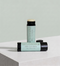 Two tubes of Tallopharm Mint Lip Balm on a white stone surface against a pale sage green background. One tube is upright and the other lies on its side, both with black caps and mint green labels.