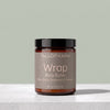A jar of Tallopharm Wrap Body Butter on a light gray stone surface against a sage green background. The product is in an amber glass jar with a black lid and a muted taupe label.