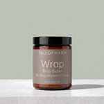 A jar of Tallopharm Wrap Body Butter on a light gray stone surface against a sage green background. The product is in an amber glass jar with a black lid and a muted taupe label.