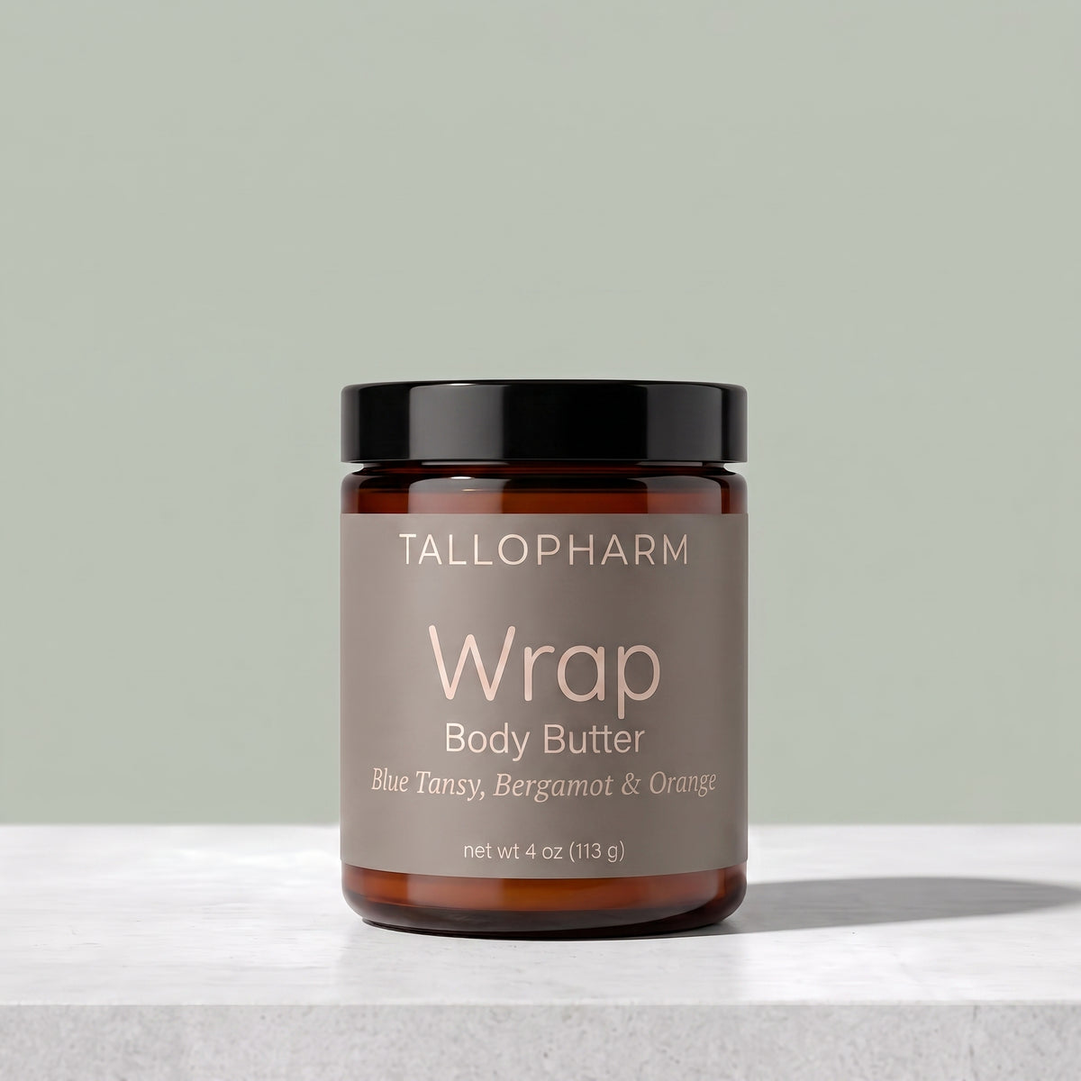 A jar of Tallopharm Wrap Body Butter on a light gray stone surface against a sage green background. The product is in an amber glass jar with a black lid and a muted taupe label.