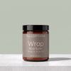 A jar of TALLOPHARM Wrap Body Butter in Mango & Bergamot scent, featuring an amber glass jar with a black lid and a muted taupe label, set on a light gray stone surface against a sage green background.
