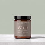 A jar of TALLOPHARM Wrap Body Butter in Mango & Bergamot scent, featuring an amber glass jar with a black lid and a muted taupe label, set on a light gray stone surface against a sage green background.