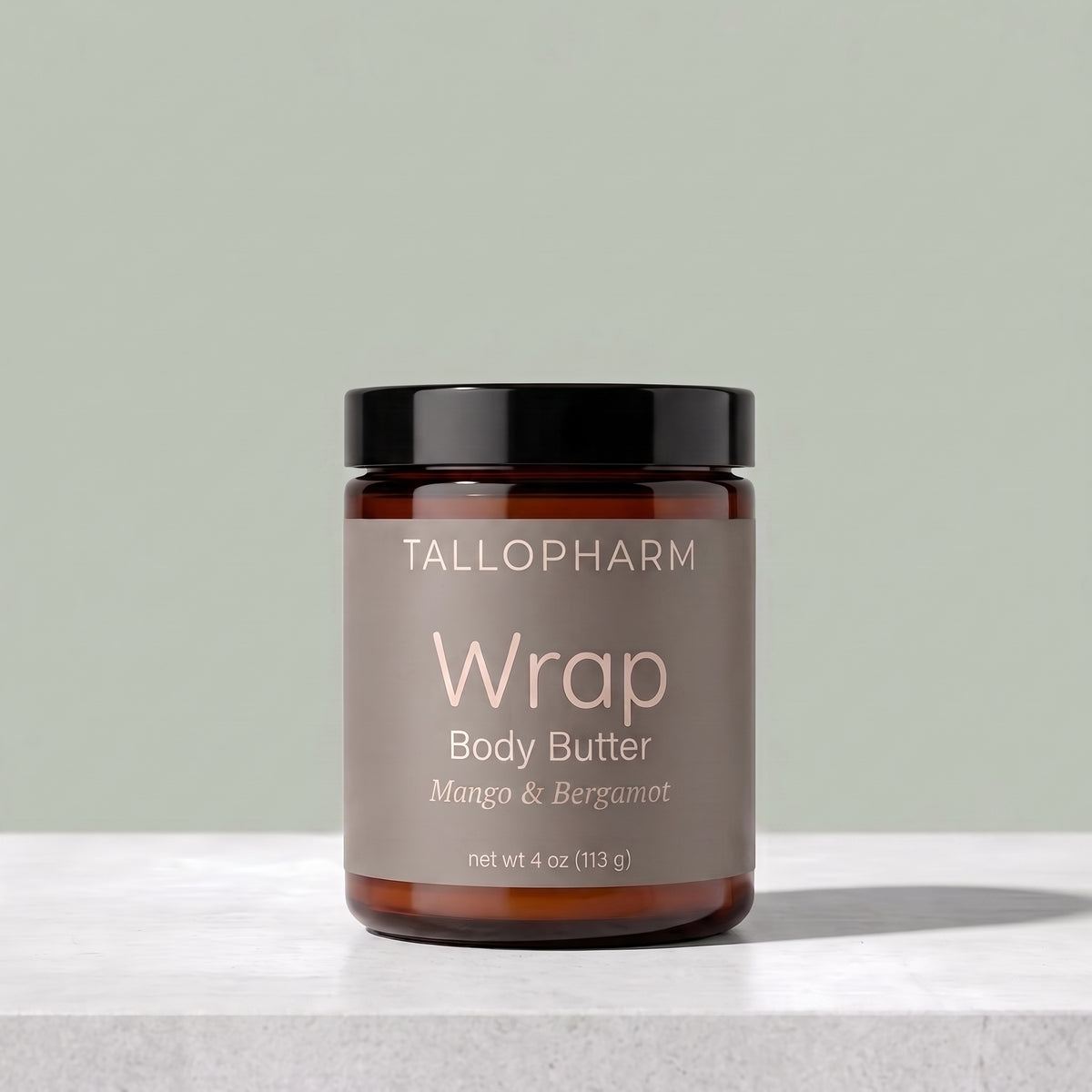 A jar of TALLOPHARM Wrap Body Butter in Mango & Bergamot scent, featuring an amber glass jar with a black lid and a muted taupe label, set on a light gray stone surface against a sage green background.