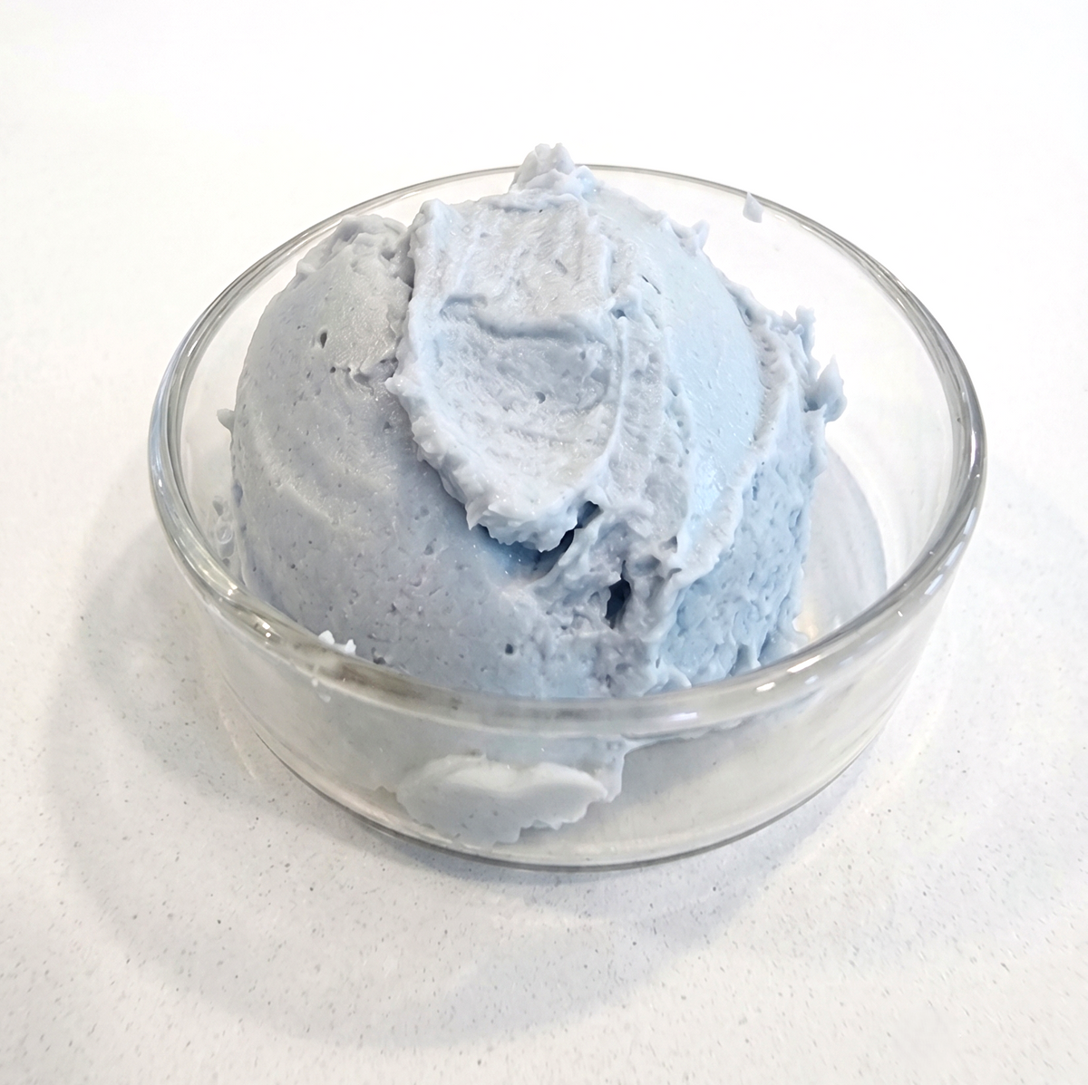 A scoop of light blue-gray whipped bergamot body butter in a small clear glass bowl, photographed on a plain light background.