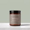 A jar of TALLOPHARM Wrap Body Butter on a light gray stone surface against a sage green background. The product is in an amber glass jar with a black lid and a taupe label.
