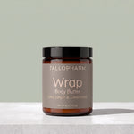 A jar of TALLOPHARM Wrap Body Butter on a light gray stone surface against a sage green background. The product is in an amber glass jar with a black lid and a taupe label.