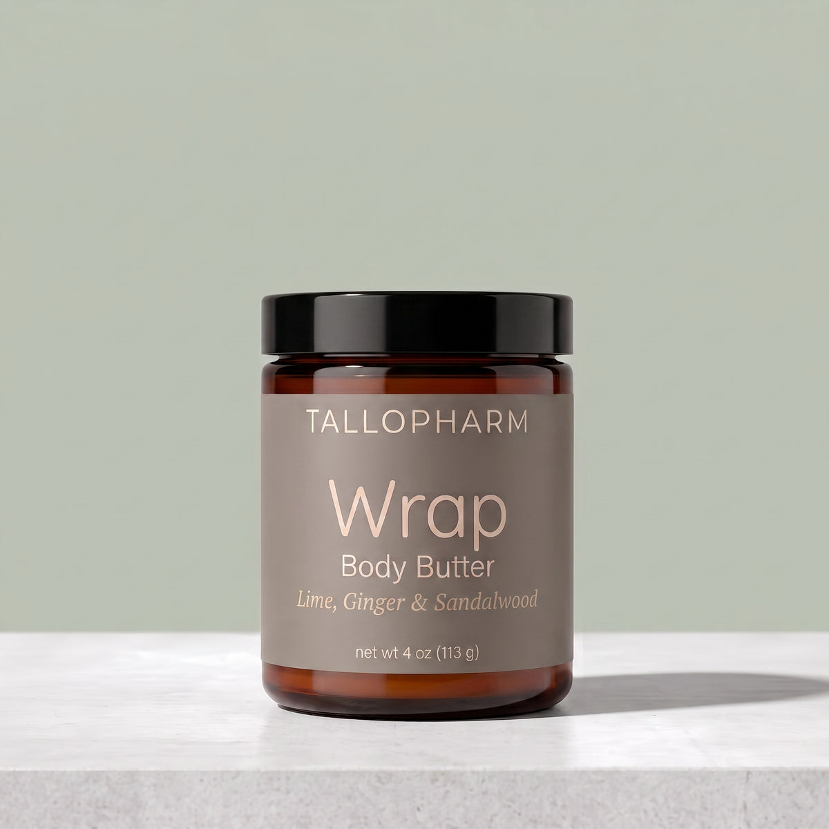 A jar of TALLOPHARM Wrap Body Butter on a light gray stone surface against a sage green background. The product is in an amber glass jar with a black lid and a taupe label.