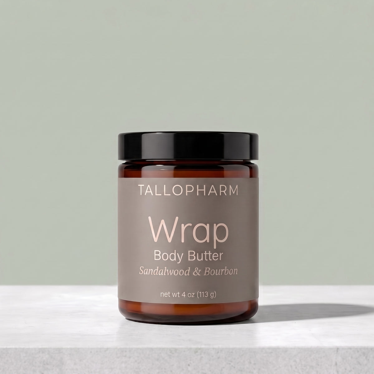 A jar of TALLOPHARM Wrap Body Butter in Sandalwood and Bourbon scent, featuring an amber glass jar with a black lid and a muted taupe label, set on a light gray stone surface against a sage green background.