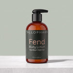 A bottle of Tallopharm Fend Body Lotion with 'Outdoor Defense' text, featuring an amber brown bottle, black pump, and a dark charcoal label, set on a light gray surface against a sage green background.