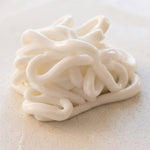 A close-up of a white, glossy, thick, and squishy lotion arranged in a loose, twisted pile on a light beige textured surface.