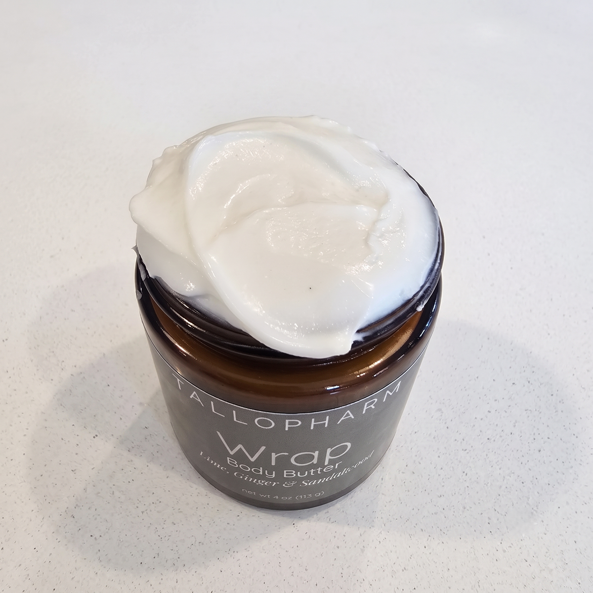 A jar of Tallopharm Wrap Lime Ginger & Sandalwood Body Butter, with a thick white cream piled on top, sits on a light, speckled countertop.
