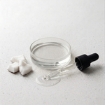 Glass dropper with black cap, small round container with coconut oil, and white coconut cubes on a light gray surface