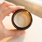 A hand with manicured nails holds an open amber glass jar filled with a white cream product, set against a warm, softly lit neutral background.