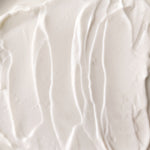 A close-up texture shot of a thick, creamy off-white cosmetic cream with soft vertical swirled lines.