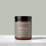 A jar of Tallopharm Wrap Body Butter in Fresh Pear, featuring an amber glass jar, a black lid, and a taupe label, set on a light gray marble surface against a sage green background.