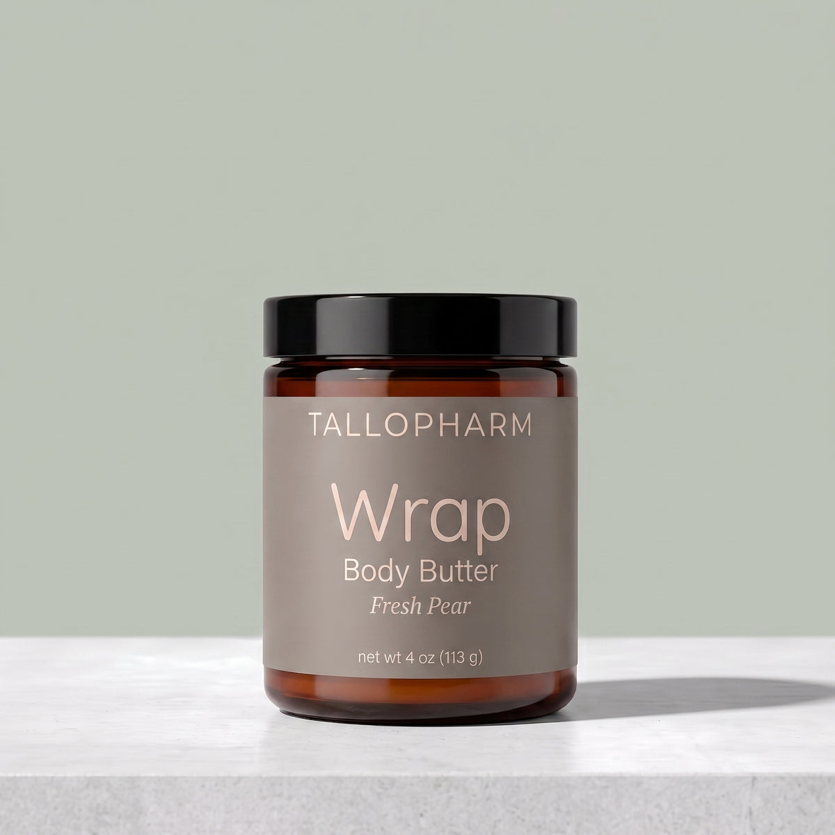 A jar of Tallopharm Wrap Body Butter in Fresh Pear, featuring an amber glass jar, a black lid, and a taupe label, set on a light gray marble surface against a sage green background.