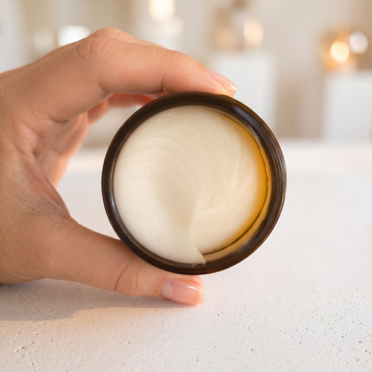 A hand holding an open round jar filled with thick, creamy off-white balm, set against a softly blurred light background.