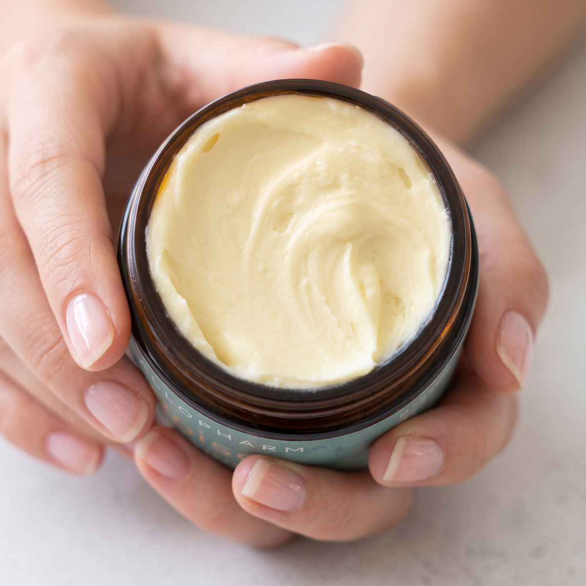 A close-up of a person's hands holding an open amber glass jar filled with pale yellow cream, with the brand name TalloPharm partially visible on the side.