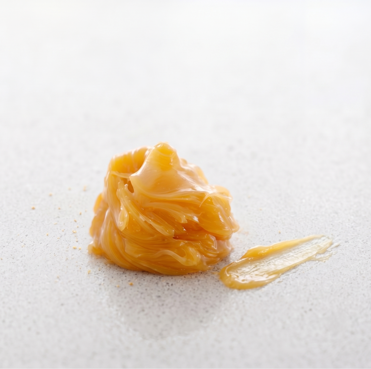 A dollop of thick, glossy golden-yellow lanolin with a small smear beside it on a light gray textured surface.