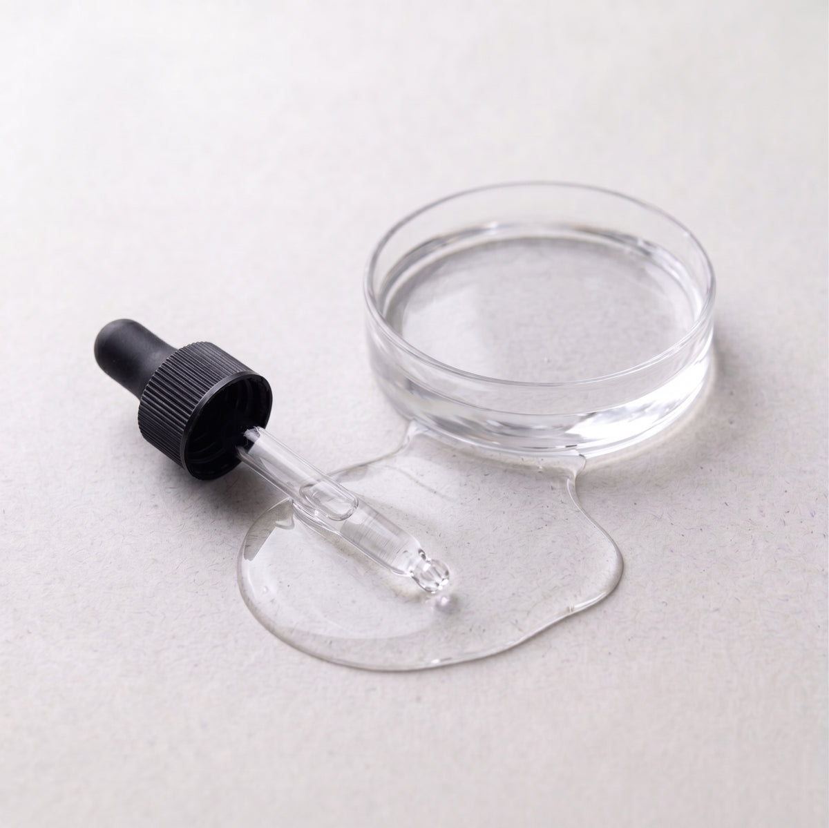 A clear cosmetic dropper bottle with a black dropper cap and a small round clear container, shown on a light gray background with Organic Vegetable Glycerin puddle in front.
