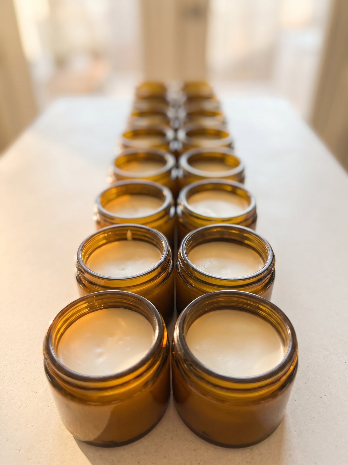 Multiple amber glass jar candles arranged in two rows on a light tabletop, photographed in warm natural light with a softly blurred indoor background.