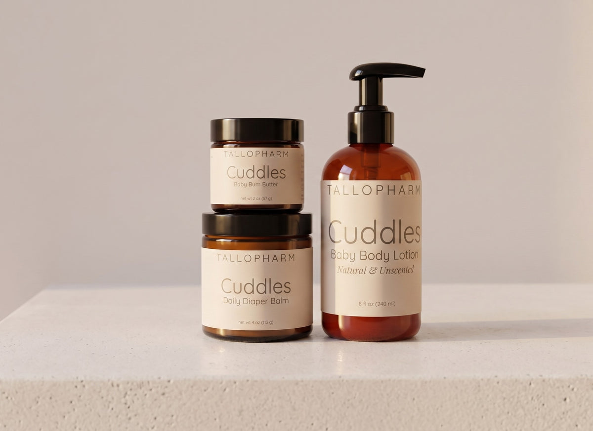 Three Tallopharm Cuddles baby care products arranged on a light stone surface against a soft beige background: two amber jars with black lids and one amber pump bottle with a black dispenser. The labels read Cuddles Daily Diaper Balm, Cuddles Baby Body Lotion, and Baby bum butter.