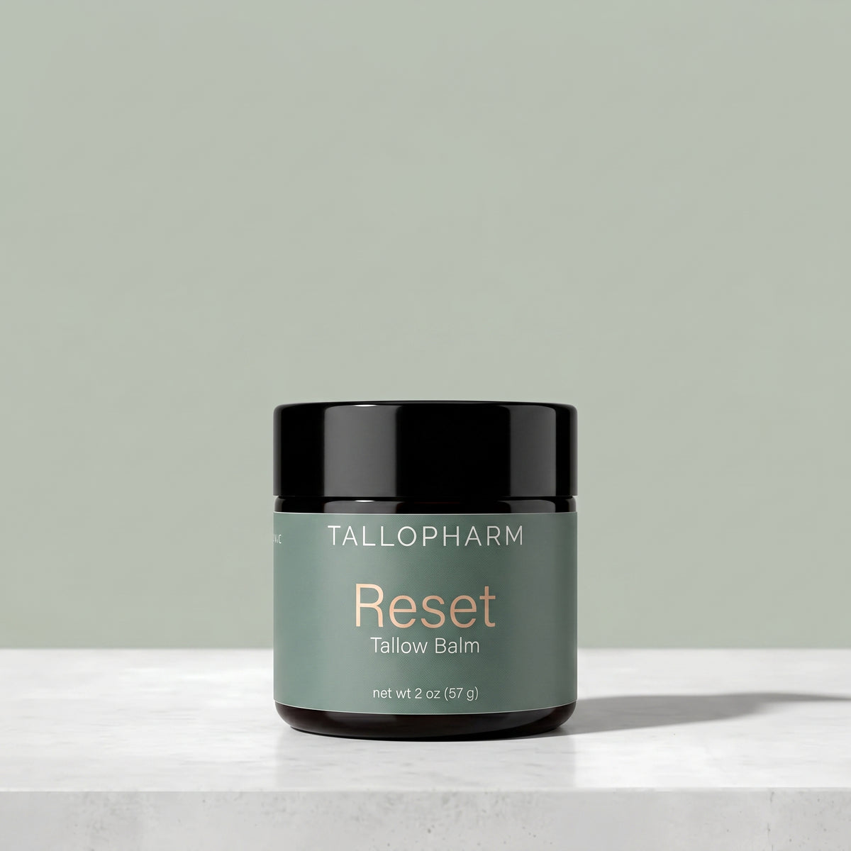 A jar of Tallopharm Reset Tallow Balm sits on a light concrete surface against a muted sage green background. The jar has a black lid and a sage green label with gold and white text.
