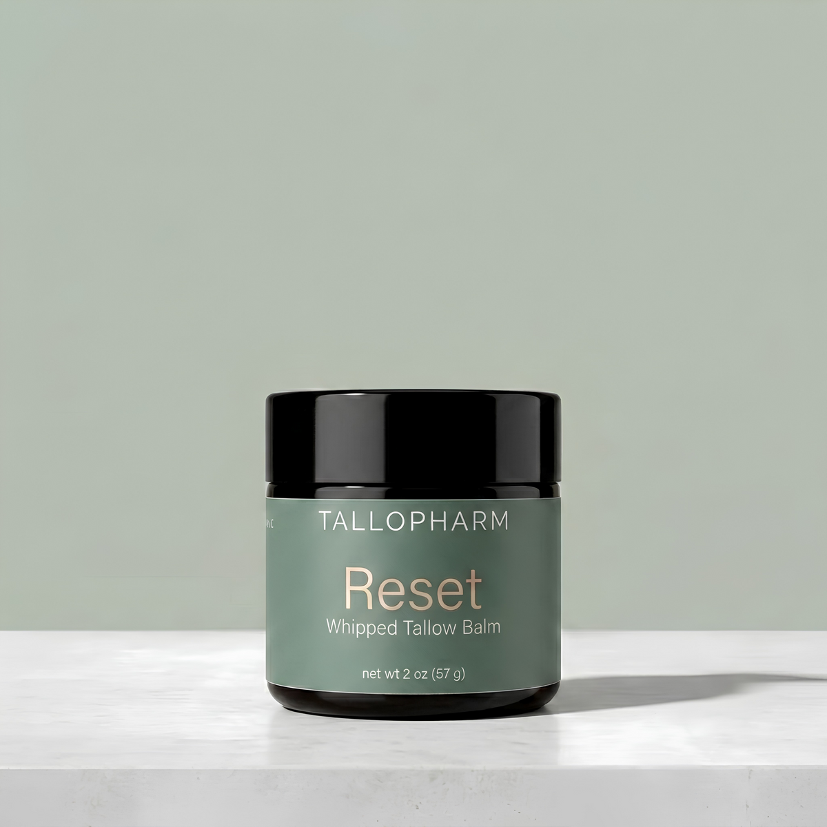 A jar of Tallopharm Reset Whipped Tallow Balm sits on a white concrete surface against a sage green background.