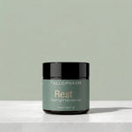 A jar of Tallopharm Rest Overnight Moisturizer with a sage green label and black lid, sitting on a white concrete surface against a sage green background.