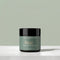 A jar of Tallopharm Rest Overnight Moisturizer with a sage green label and black lid, sitting on a white concrete surface against a sage green background.