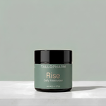 A jar of TALLOPHARM Rise Daily Moisturizer with a sage green label and black lid, sitting on a white concrete surface against a matching sage green background.