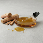 Glass dropper with amber liquid next to sandal wood sticks on a light gray background