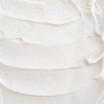 A close-up texture shot of a thick, creamy off-white cosmetic cream or lotion swirled on a white surface.