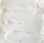 A close-up texture shot of a thick, creamy off-white cream, showing smooth horizontal swaths and a glossy, slightly translucent surface.